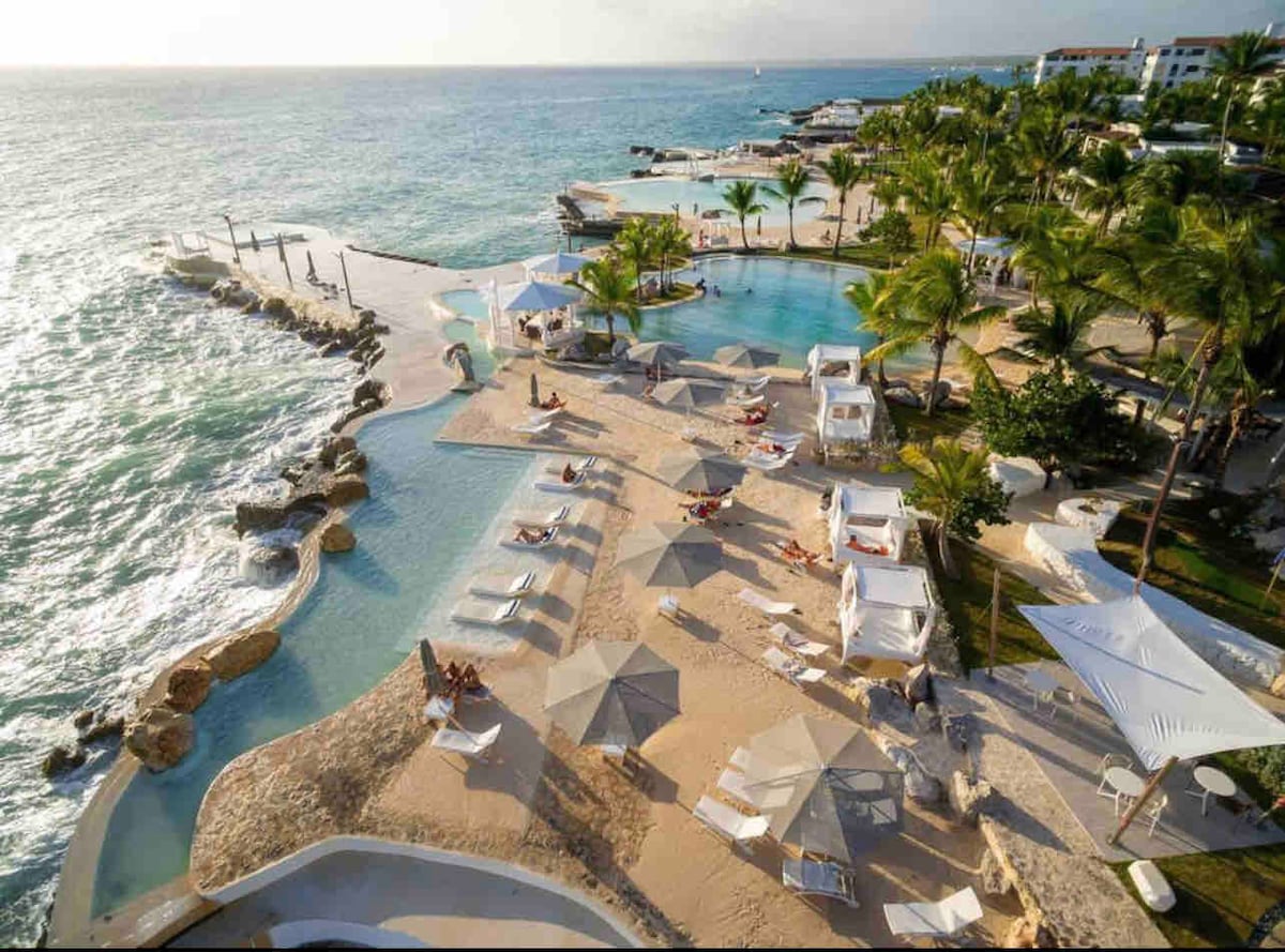 The image shows a spacious beachfront area featuring multiple pools surrounded by sandy areas. Lounge chairs and shaded cabanas are placed strategically, offering ample relaxation spaces. Palm trees frame the scene, with gentle waves visible in the background, contributing to a serene atmosphere.