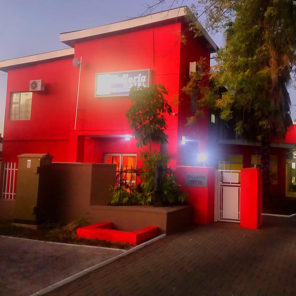 The exterior of Galleria Hotel features a vibrant red facade, complemented by white signage. Lush greenery and landscaping adorn the entrance, while warm lights illuminate the surroundings. The structure is set within a well-maintained pathway, enhancing access to the entrance area.