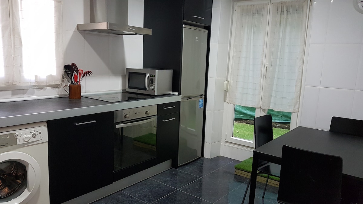 The kitchen features modern appliances including a refrigerator and microwave, set against sleek black cabinets. A dining table with four black chairs is positioned near a window, allowing natural light to brighten the space. A washing machine is located under the countertop.