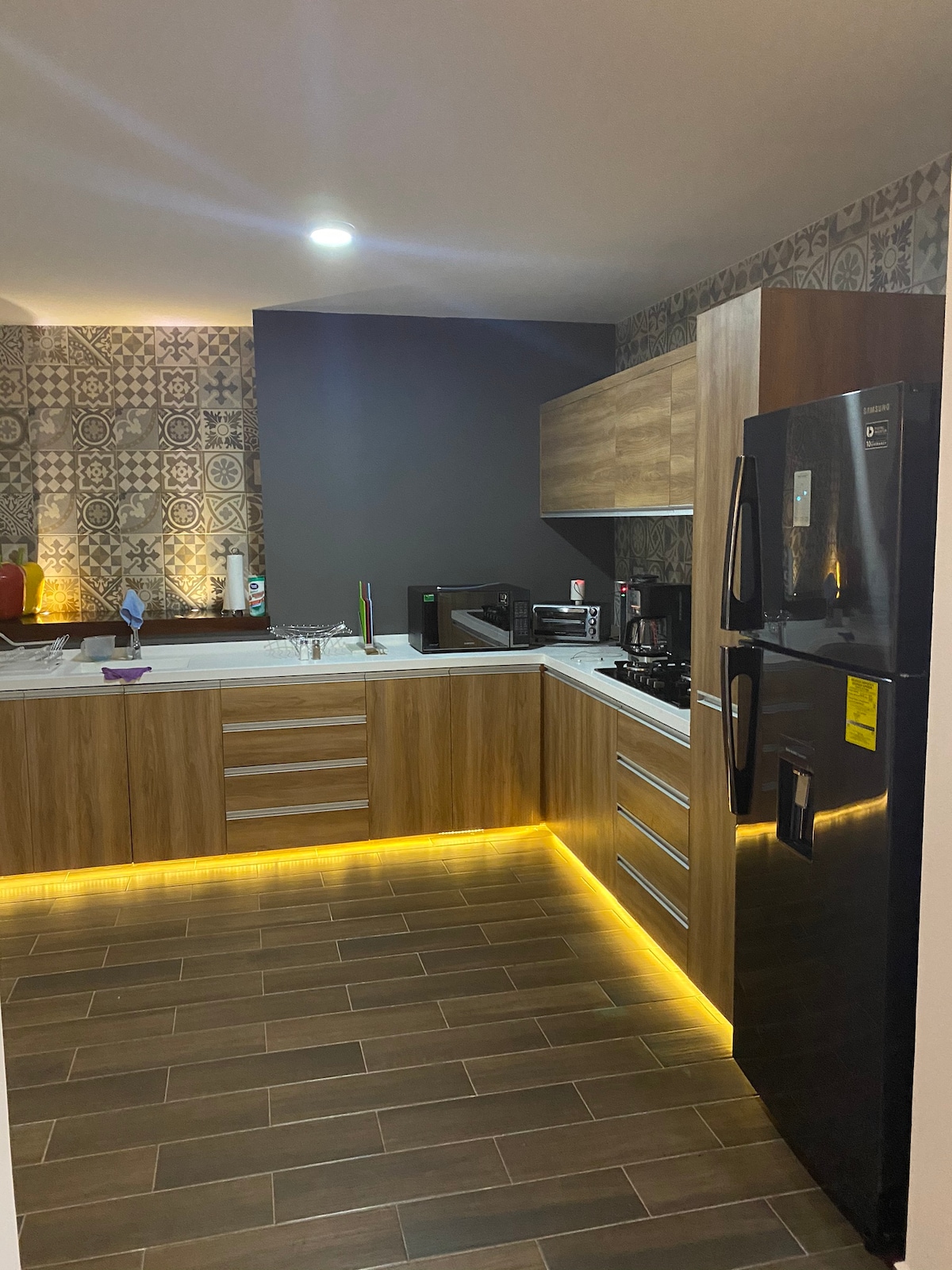 A modern kitchen features wooden cabinetry and a sleek black refrigerator. Under-cabinet lighting adds warmth to the space, highlighting the countertops and patterned wall. Kitchen appliances, including a microwave and stovetop, are conveniently arranged for easy access.