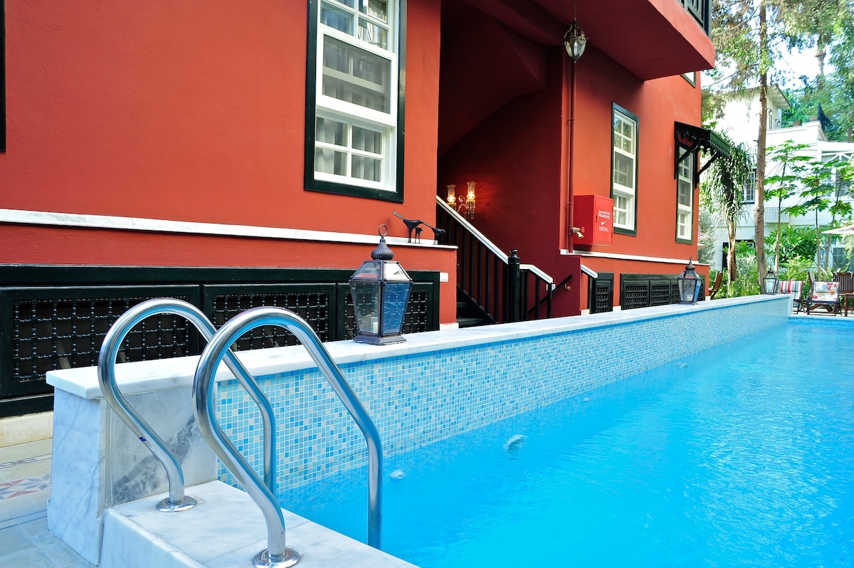 A modern pool is framed by a red building and features silver handrails. The water is clear and inviting, reflecting the surrounding greenery and light. Lantern-style lighting is seen nearby, adding a subtle decorative touch to the pool area.