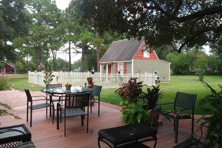 A spacious outdoor patio area features a round dining table surrounded by chairs, set against a backdrop of lush greenery. A cozy cottage with red accents and a white picket fence is visible in the distance, providing a serene country setting.