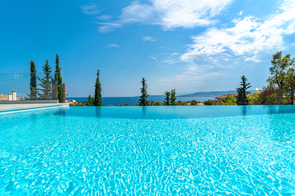 An expansive infinity pool reflects clear blue waters, set against a backdrop of lush greenery. The shimmering surface meets the horizon, where the sea meets the sky, creating a seamless view that captures the serene landscape.