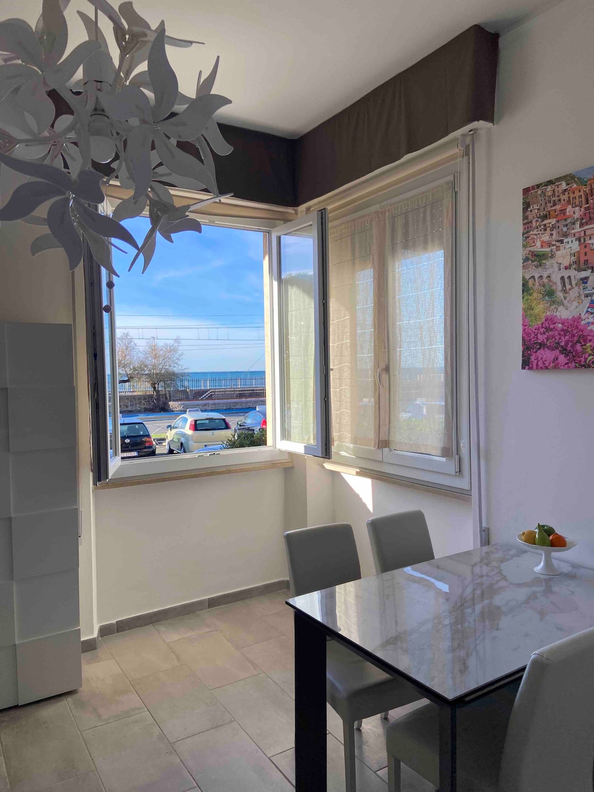 A light-filled dining area features a table with four chairs and a bowl of fruit. Large windows open to scenic views, allowing natural light to enhance the space. A colorful artwork adorns the wall, contributing to the room's inviting atmosphere.