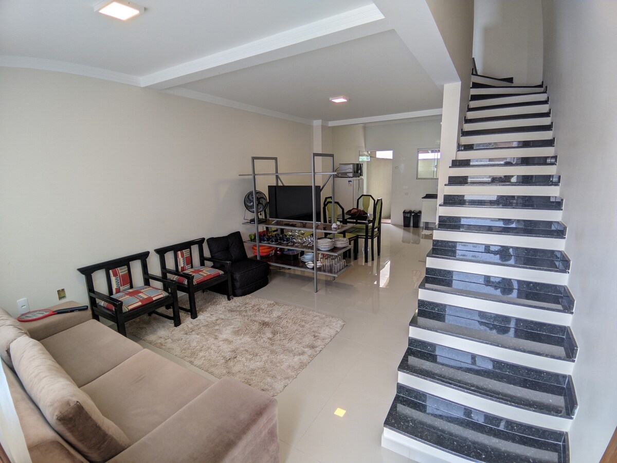 The living area showcases a light-toned sectional sofa alongside black armchairs with colorful cushions. A large TV is positioned against the wall, while a dining area is visible in the background. The space is complemented by glossy tiled flooring and a staircase leading to the upper level.