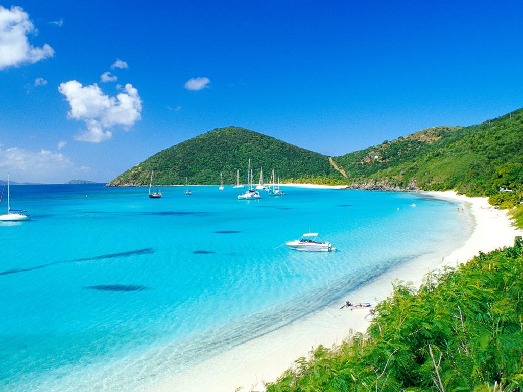 A tranquil beach scene is depicted, showcasing soft white sand and crystal-clear turquoise waters. Gentle waves lap against the shore, with a few sailboats anchored in the distance. Lush greenery covers the hills in the background under a clear blue sky, creating a serene coastal landscape.