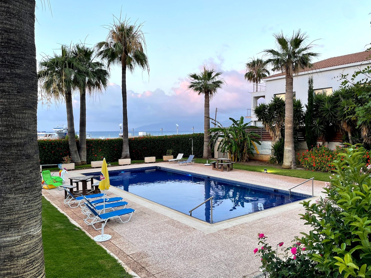 A swimming pool is surrounded by palm trees and manicured gardens. Lounge chairs and a small table are positioned near the water, providing a space for relaxation. Umbrellas offer shade, while vibrant flowers add color to the landscape.