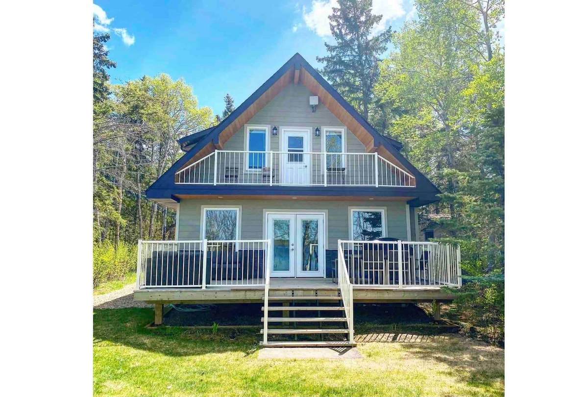 A two-story cottage is set amidst lush greenery, featuring a large front deck with white railings. Steps lead up to the entrance, which is flanked by multiple large glass doors that allow natural light to fill the interior.