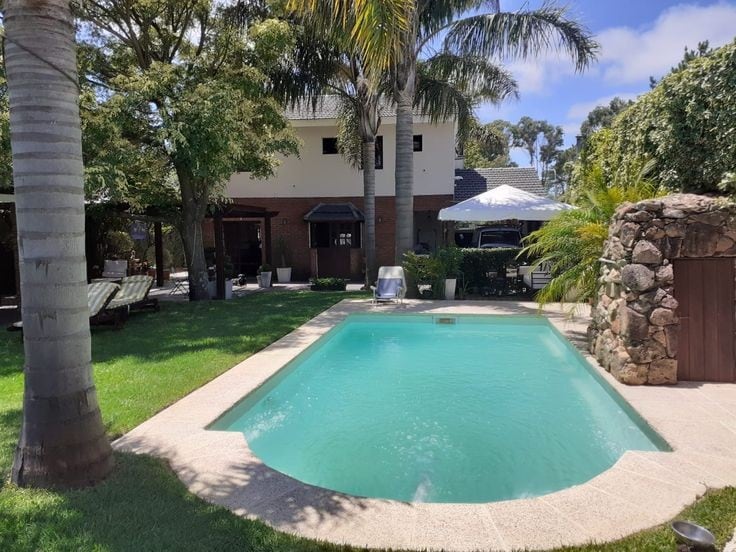 A refreshing swimming pool is positioned in a lush garden, surrounded by palms and greenery. Lounge chairs are available for relaxation. A stone outdoor grill and shaded areas provide options for gathering. The overall space delivers a serene ambiance for guests.