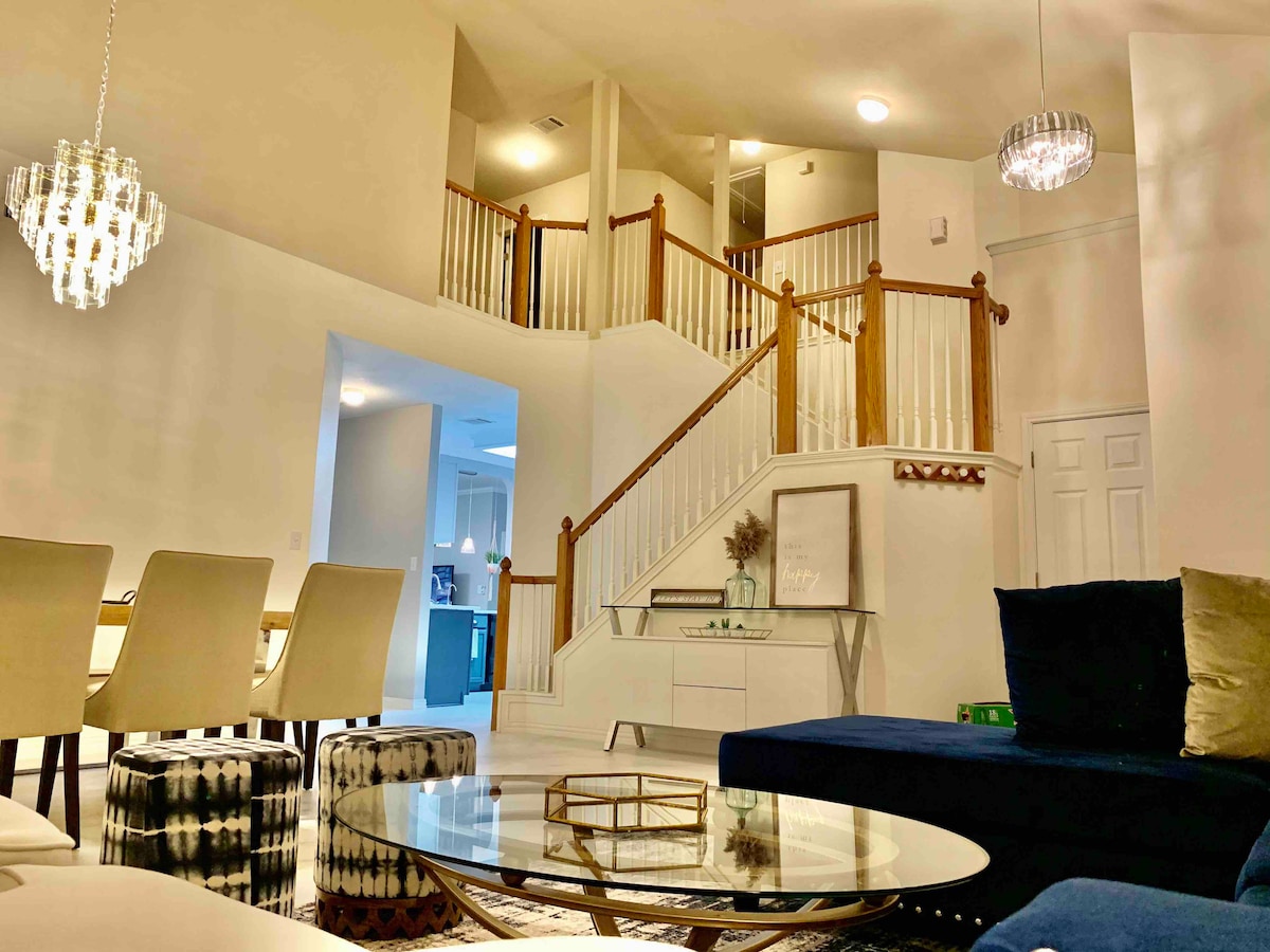 A spacious living area features a large sectional sofa and a round coffee table. Natural light floods the space from multiple levels, highlighting the elegant staircase with wooden railings. A dining area with light-colored chairs is visible in the background, contributing to an open and airy environment.