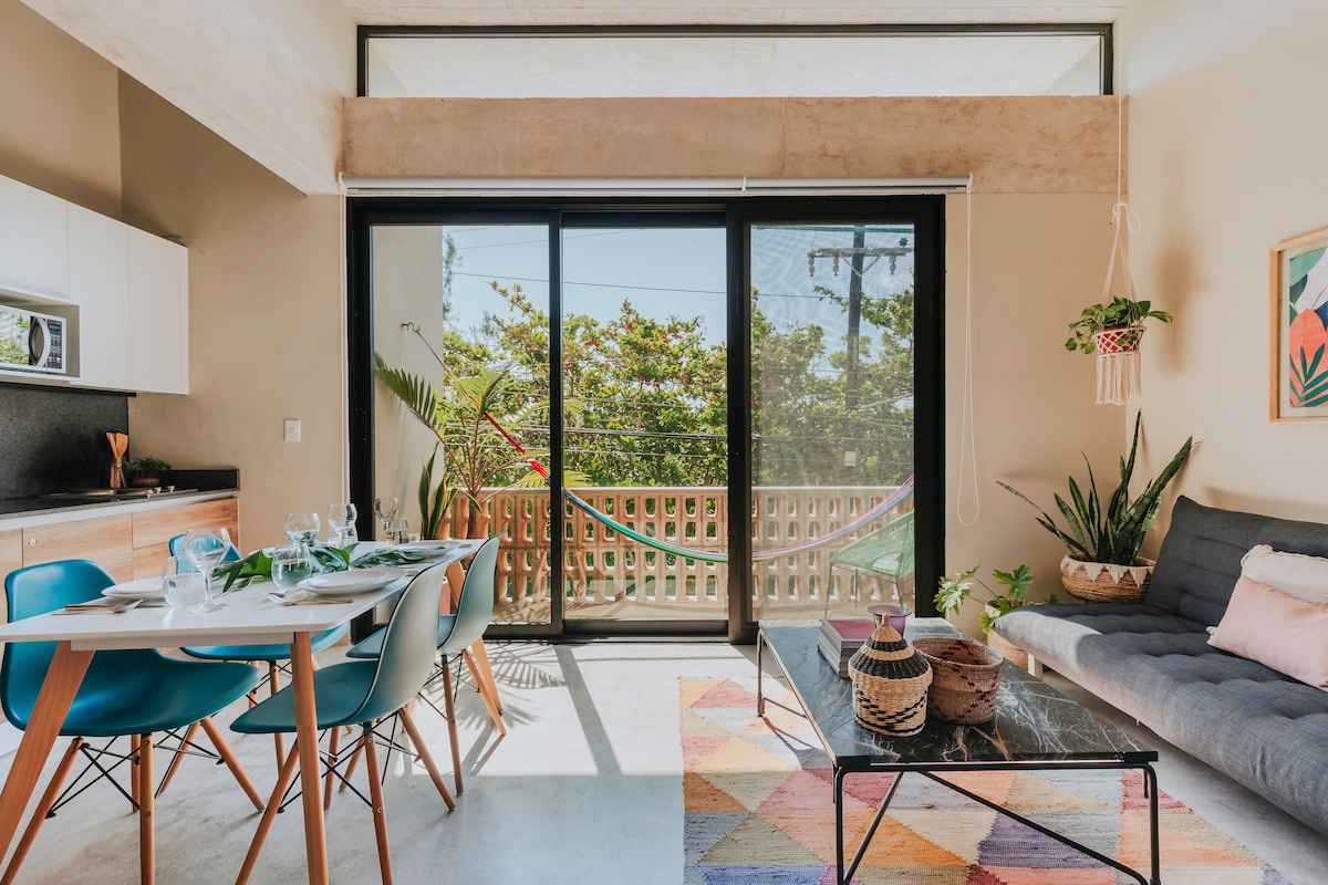 A modern living area features large glass sliding doors that open to a private balcony with a hammock. A dining table with blue chairs is positioned near the kitchen, while a sofa and colorful area rug add comfort and style to the space.