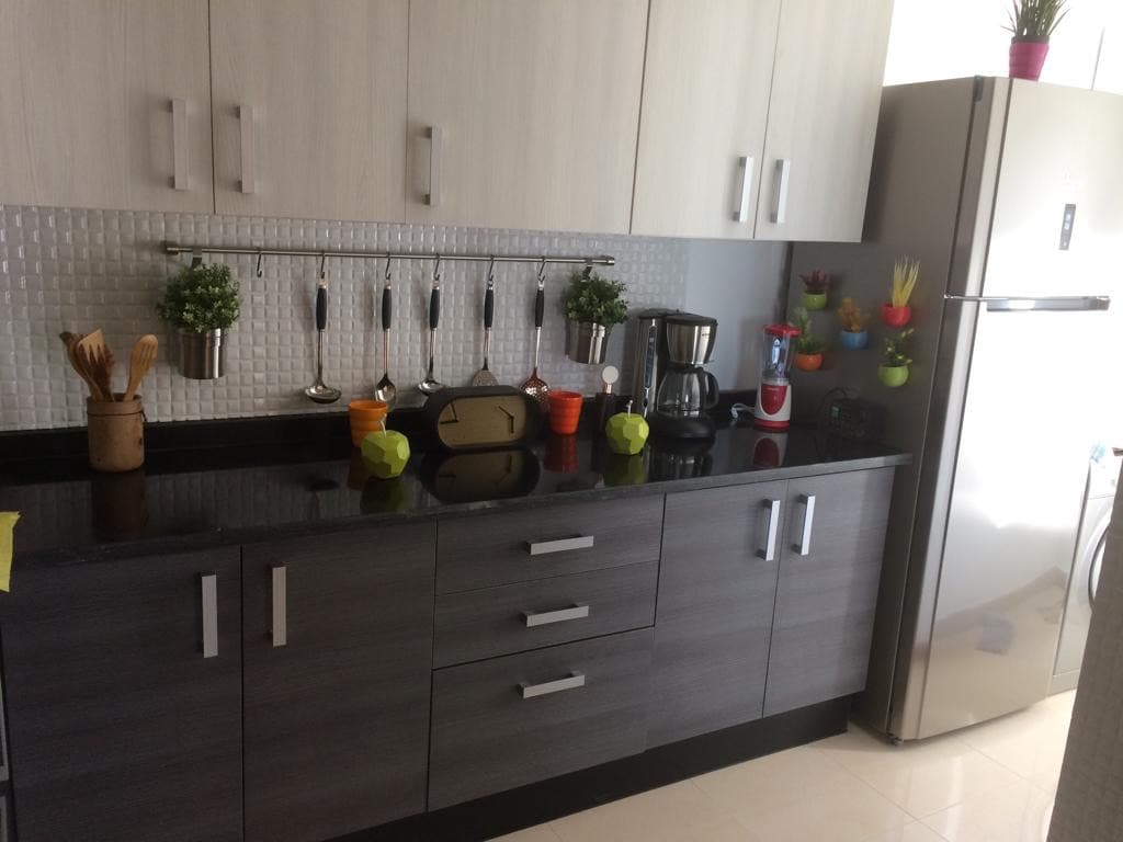 A modern kitchen features a sleek black countertop and light wood cabinetry. Hanging utensils are organized on a rod, with colorful potted plants adding a pop of color. A stainless steel refrigerator stands next to a coffee maker and a sleek kitchen clock, emphasizing functionality.