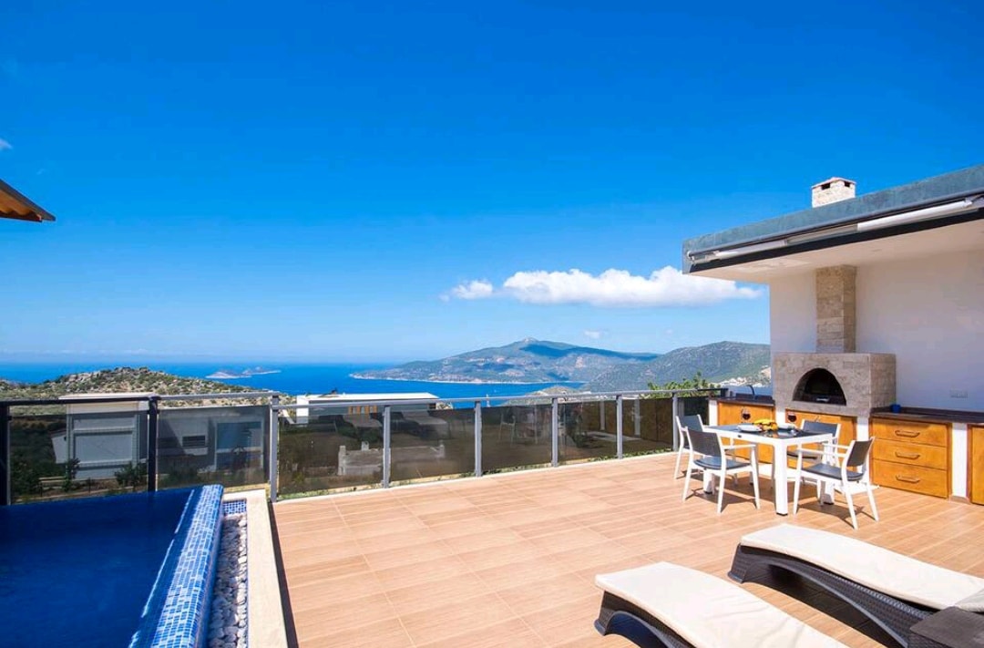 An outdoor terrace provides a scenic view of the sea and surrounding hills. A dining area features a table with seating for four, accompanied by two lounge chairs near an infinity pool. A barbecue area is visible, with clear blue skies overhead.