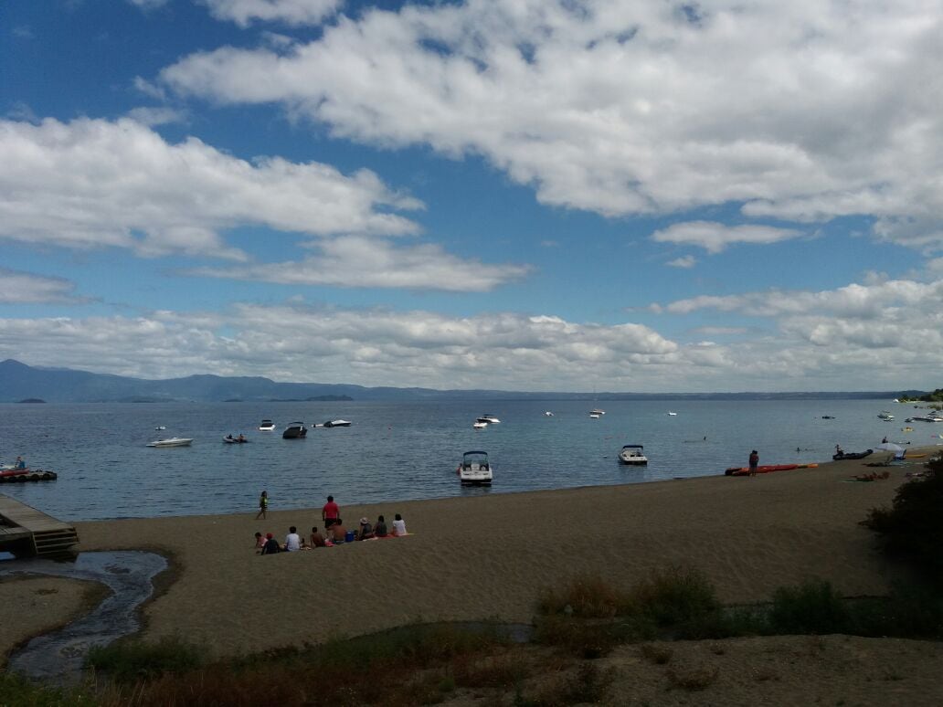 A sandy beach is presented, with calm waters dotted by various boats. People are gathered along the shoreline, enjoying the open space, while clouds drift across a blue sky. The scenic view reveals distant mountains, enhancing the serene atmosphere of the location.