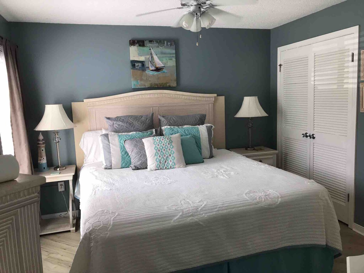 A calming bedroom features a spacious bed adorned with a light-colored quilt and accent pillows in soft blue and gray hues. Two bedside lamps provide illumination, complemented by artwork of a sailboat above the headboard. Light streams in through a window, enhancing the serene atmosphere.
