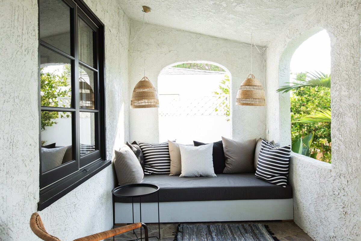 A bright and airy seating area features a built-in lounge with plush cushions in various shades, accented by a striped pattern. Two woven pendant lights hang overhead, and a small black table sits beside the lounge, providing a cozy spot for relaxation and enjoyment of the surrounding greenery.