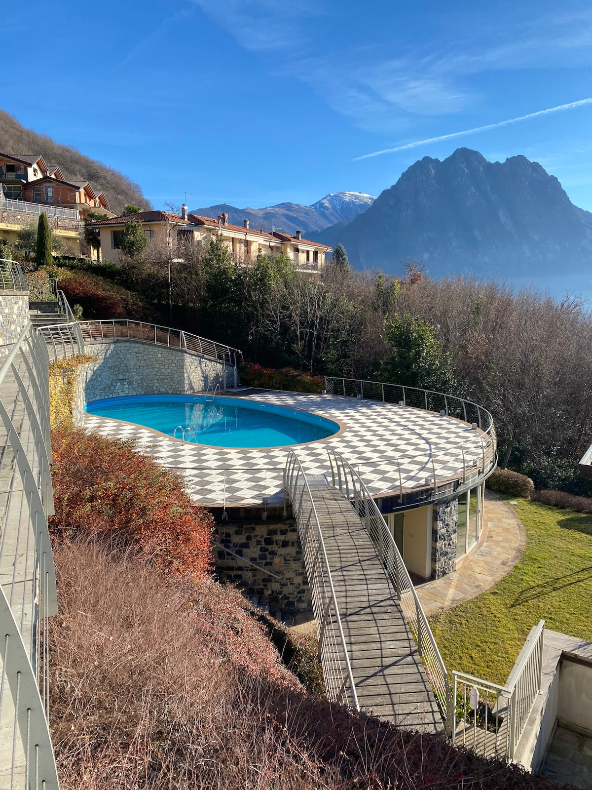 An outdoor pool with a circular shape is surrounded by a patterned terrace. Lush greenery and residential buildings are visible in the background, with mountains rising majestically under a clear blue sky.