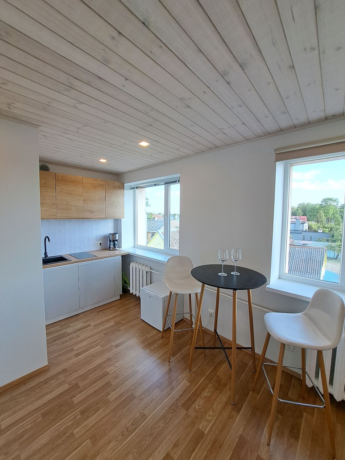 A bright kitchen area is equipped with modern cabinetry and a countertop. A small round table with two white chairs sits beside a window that offers a view of the surrounding neighborhood. Natural light enhances the warm wooden flooring.