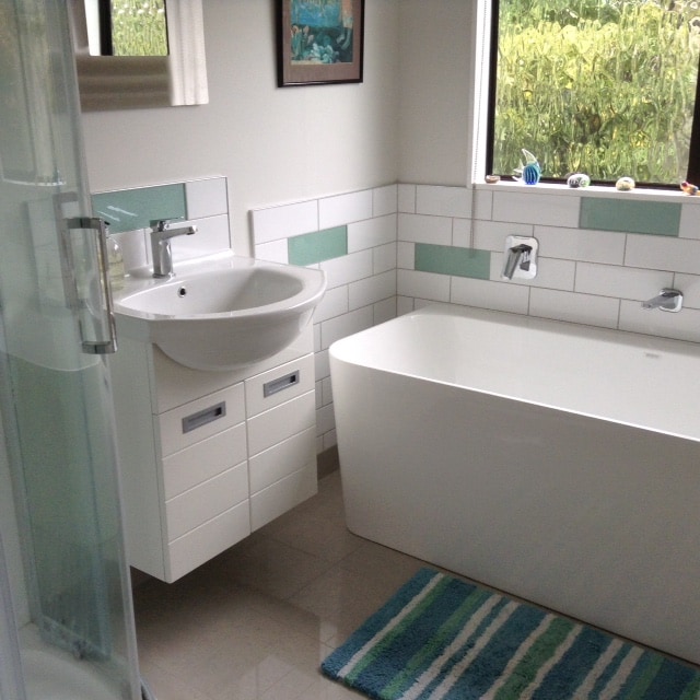 The modern bathroom features a sleek bathtub and a stylish sink with storage underneath. A glass shower enclosure is visible, along with a small window allowing natural light. The tile design includes muted color accents, and a soft rug lies on the floor.