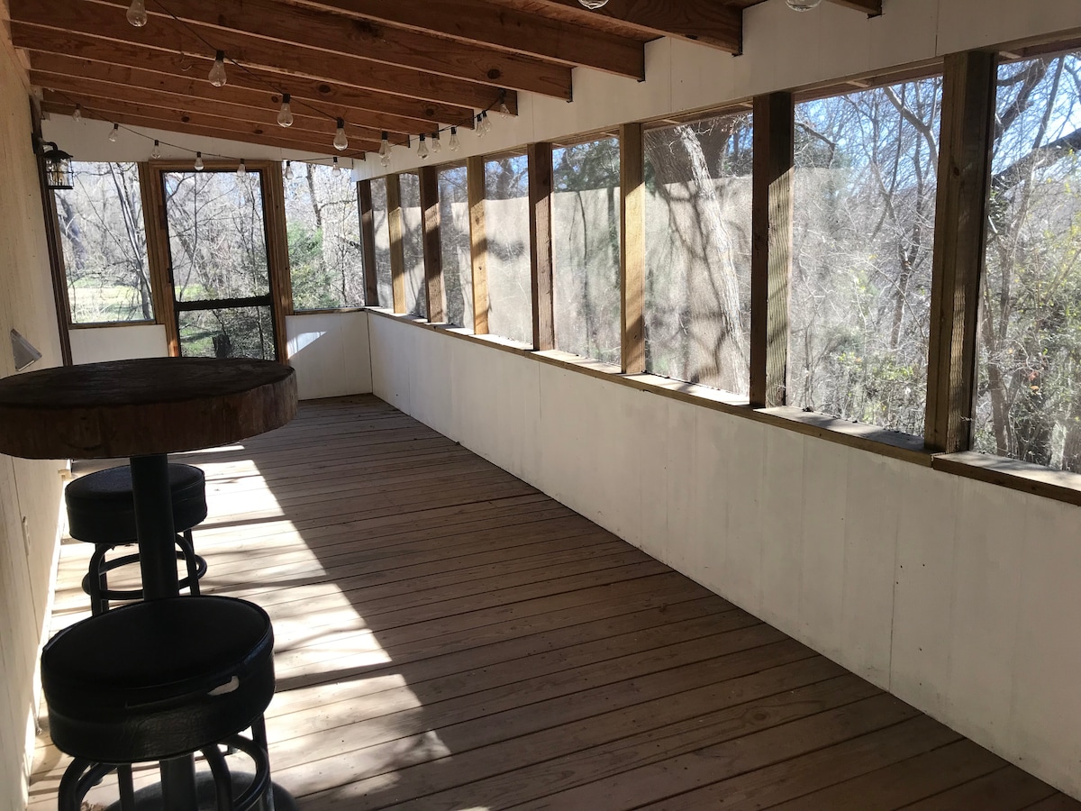 A spacious covered porch is featured with wooden beams and large windows allowing natural light to filter through. Bar stools are arranged at a circular table, providing a comfortable spot to relax while enjoying views of the surrounding nature.