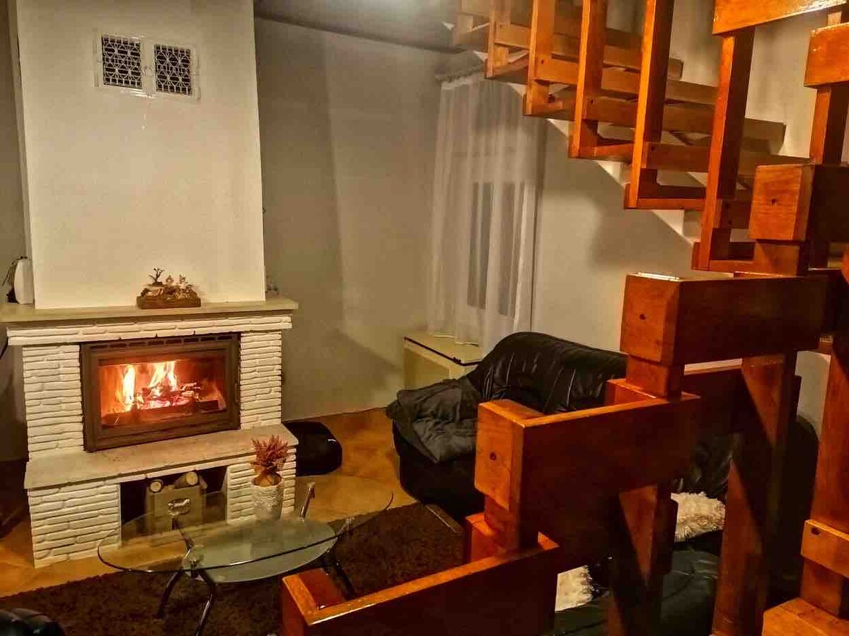 A living room is featured with a cozy fireplace, radiating warmth. Black seating arrangements are positioned around a glass coffee table. A wooden staircase leads to the upper level, complimented by natural light entering through the window, partially covered by sheer curtains.
