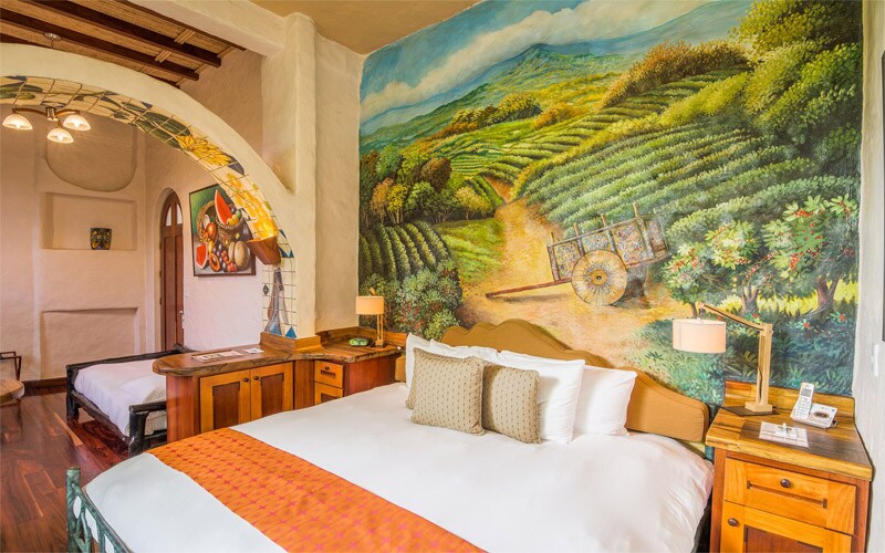 A spacious guest room features a large king-size bed with decorative pillows. A mural depicting rolling green hills and a cart is painted on one wall. A small sofa bed is positioned nearby, and wooden furniture with bedside lamps completes the inviting space.