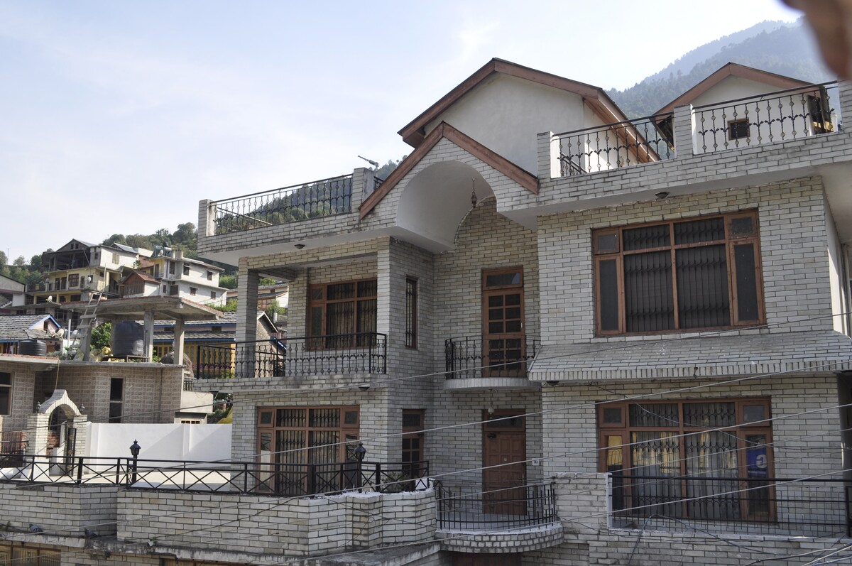 The cottage features a well-structured facade with a combination of brick and stone elements. Balconies extend from the upper level, offering views of the surrounding mountains. The building is set against a backdrop of greenery, adding to the visual appeal of the area.