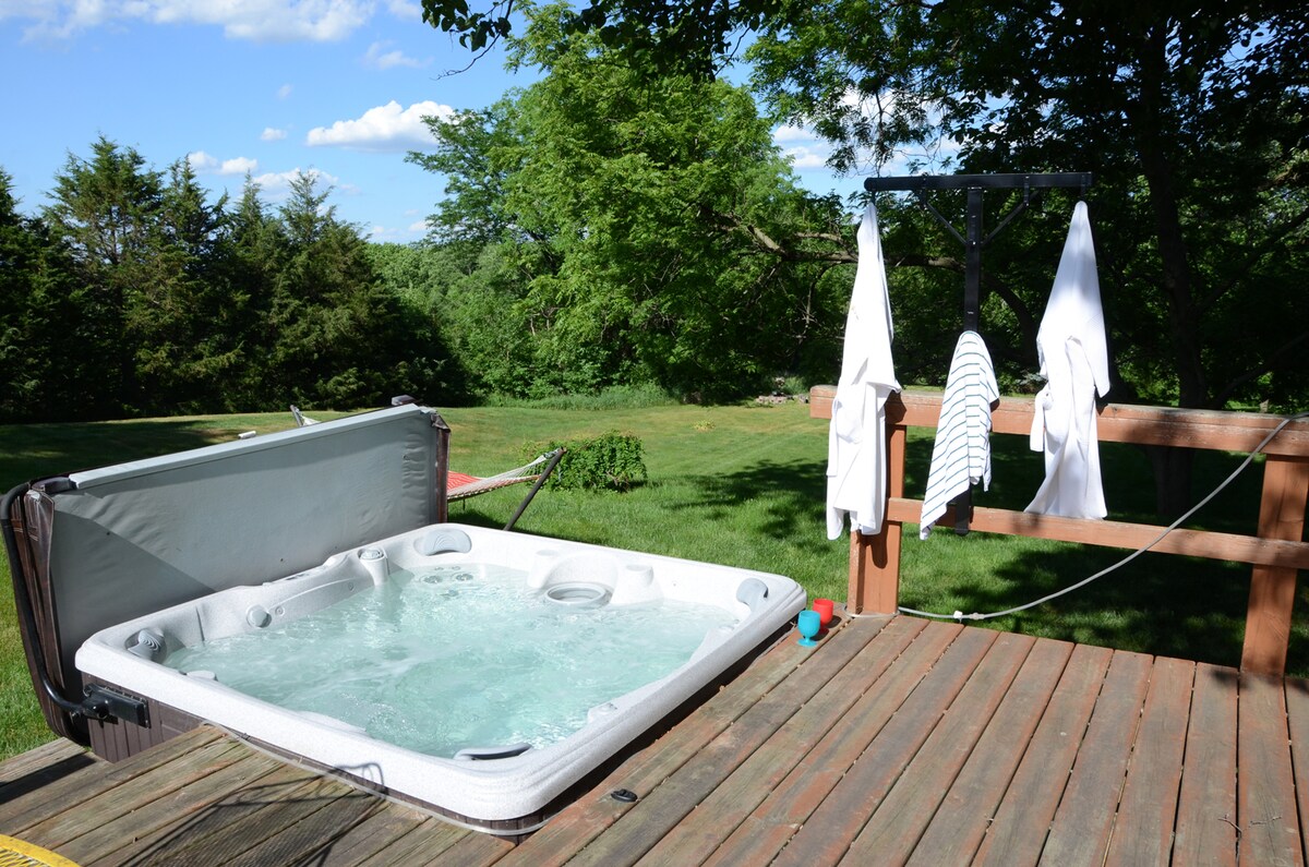 A private hot tub is situated on a wooden deck, featuring bubbling water and a retractable cover. Nearby, two towels hang on a rack, while natural greenery and an open lawn are visible in the background, providing a serene outdoor setting.