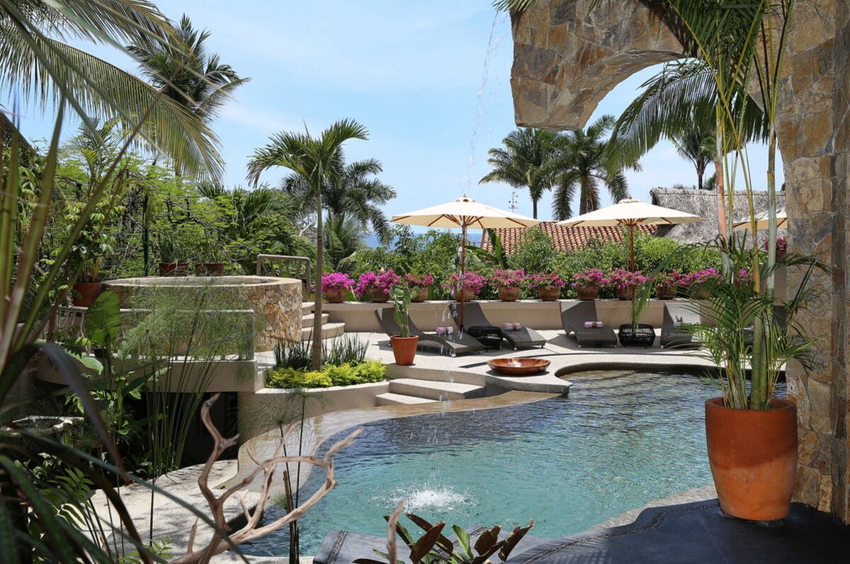 A tranquil outdoor area features a saltwater pool surrounded by lush tropical plants and flowering bougainvillea. Comfortable lounge chairs with umbrellas create a relaxing atmosphere. The flowing water of a fountain adds a serene sound, while palm trees sway against a clear blue sky.