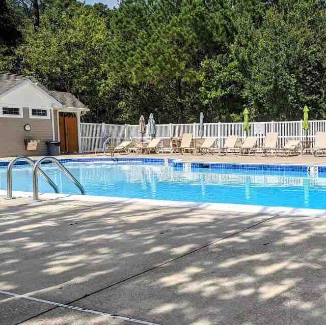 A community pool area is depicted, featuring a clear blue pool surrounded by lounge chairs. Umbrellas provide shaded seating options. Lush greenery is visible in the background, along with a poolhouse that offers amenities for guests.