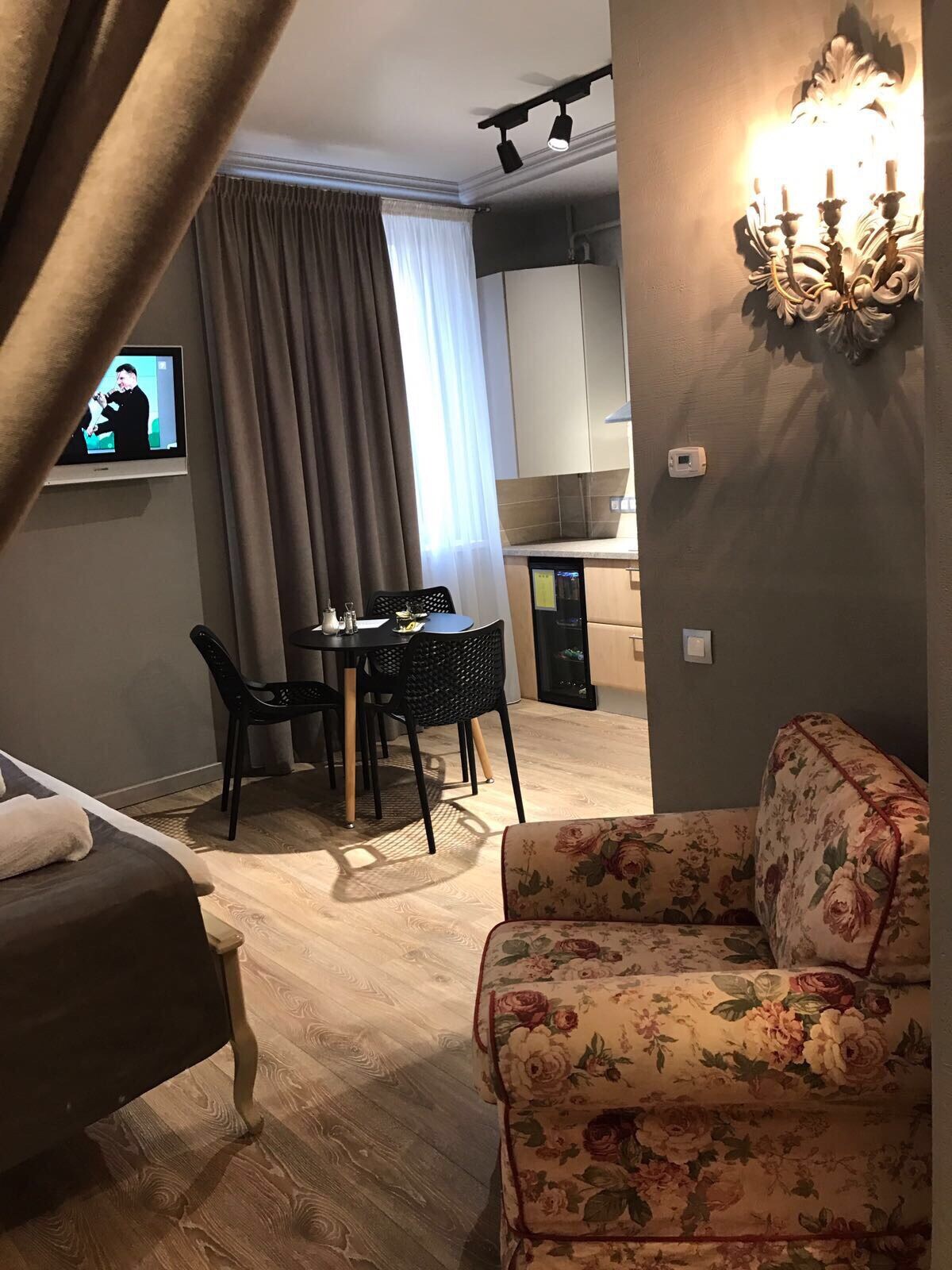 A cozy seating area includes a patterned armchair, positioned near a dining table with chairs. A wall-mounted television is visible alongside a kitchenette, while soft curtains frame a bright window, allowing natural light to fill the space.