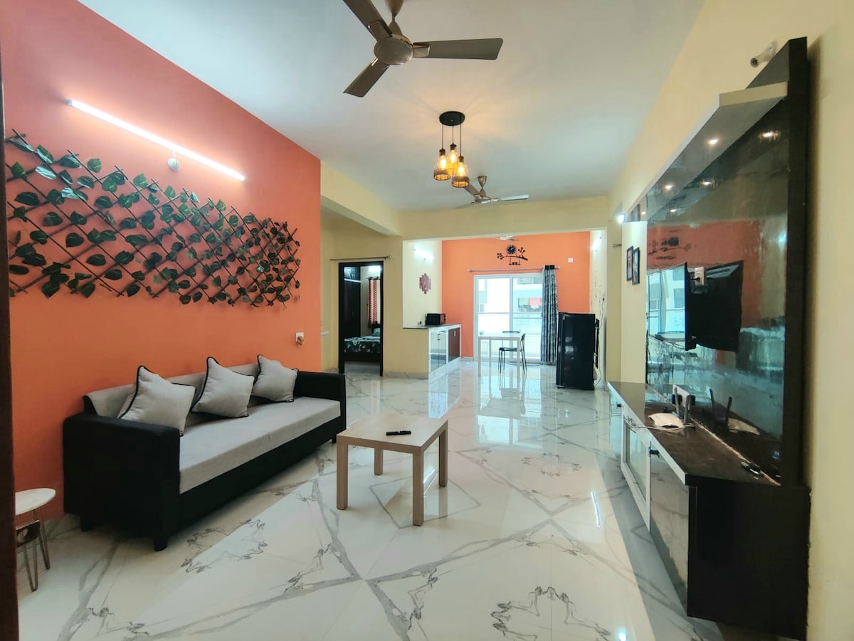 A spacious living area features a black and white sofa facing a sleek television unit with a dark finish. A light-colored, minimalist coffee table is centered on the marble floor, which reflects natural light. Wall decor includes an artistic green leaf arrangement against an orange accent wall.
