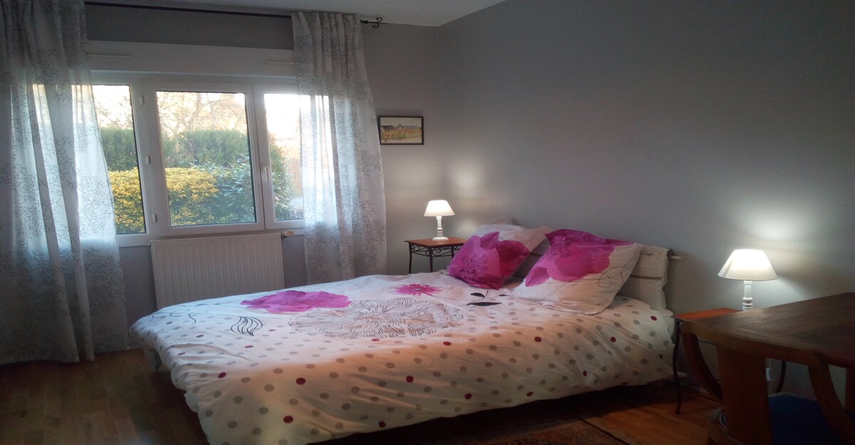 A calm bedroom features a spacious bed adorned with a patterned duvet and decorative pillows. Natural light enters through a large window, complemented by sheer curtains. A small table with a lamp is positioned next to the bed, enhancing the room's functionality.