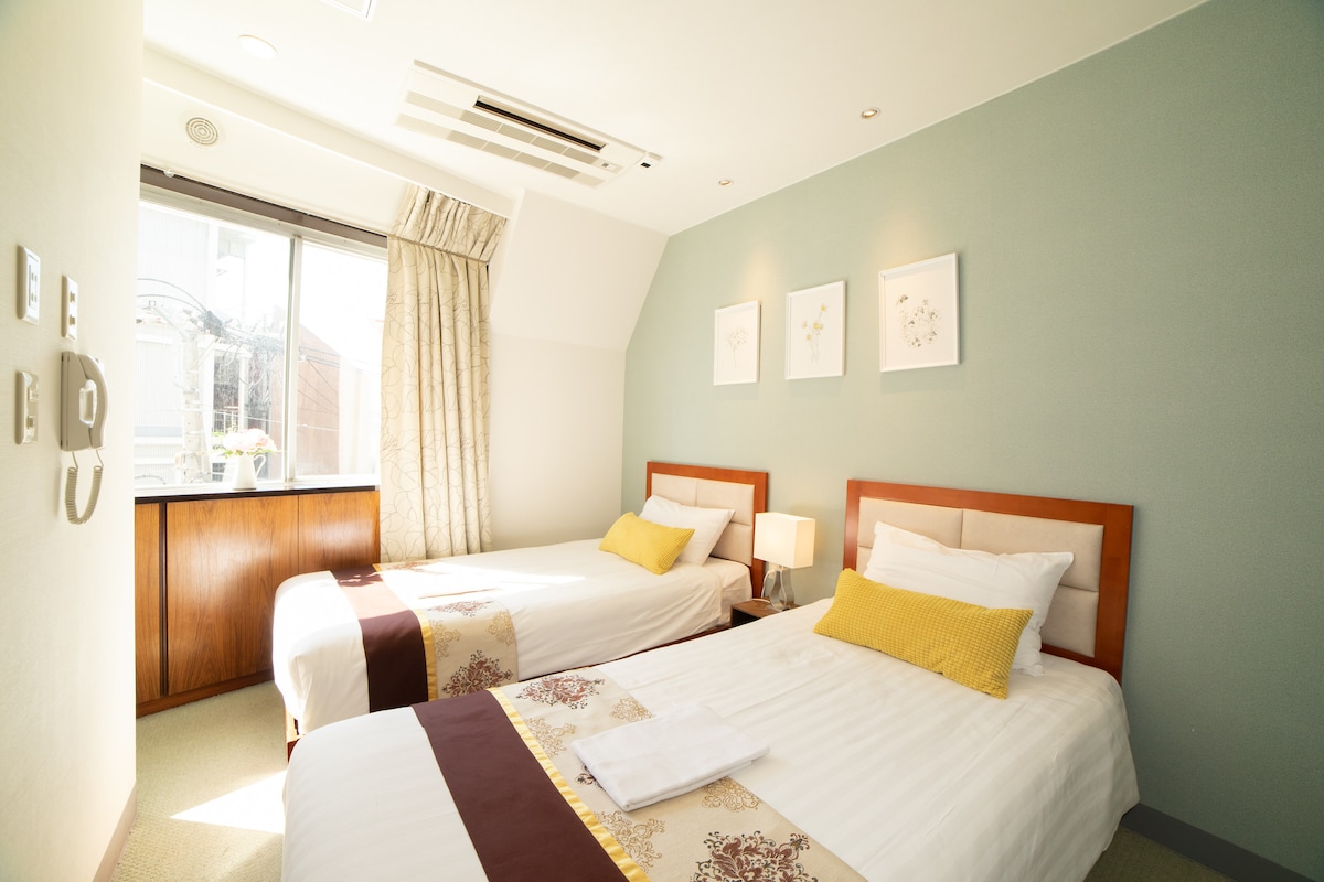 A guest room features two neatly arranged beds, each with white linens and decorative throws. A vibrant yellow pillow adds a pop of color. Natural light flows through the window, complemented by soft curtains, while framed artwork adorns the wall.