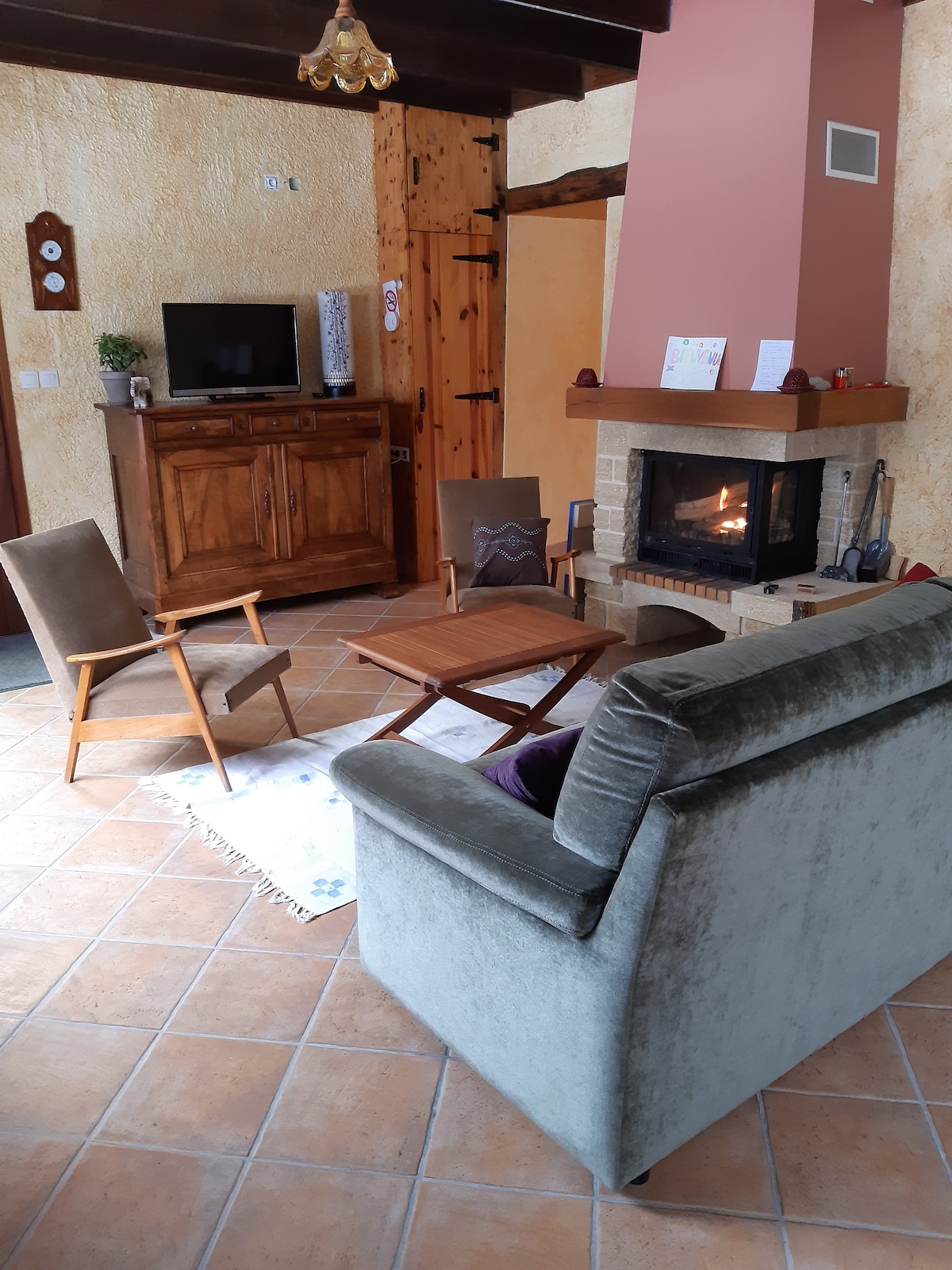 A cozy living area features a comfortable gray sofa and two wooden chairs, arranged around a small coffee table. A decorative fireplace is set against a textured wall, while a wooden cabinet stands to the side, complemented by warm lighting.