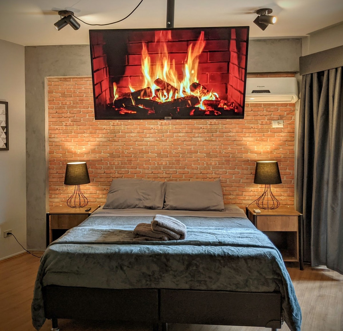 A cozy bedroom is highlighted by a feature wall of exposed brick. A large television displays a visual of a crackling fire above a spacious bed dressed in soft linens. Two nightstands with lamps flank the bed, providing additional lighting.