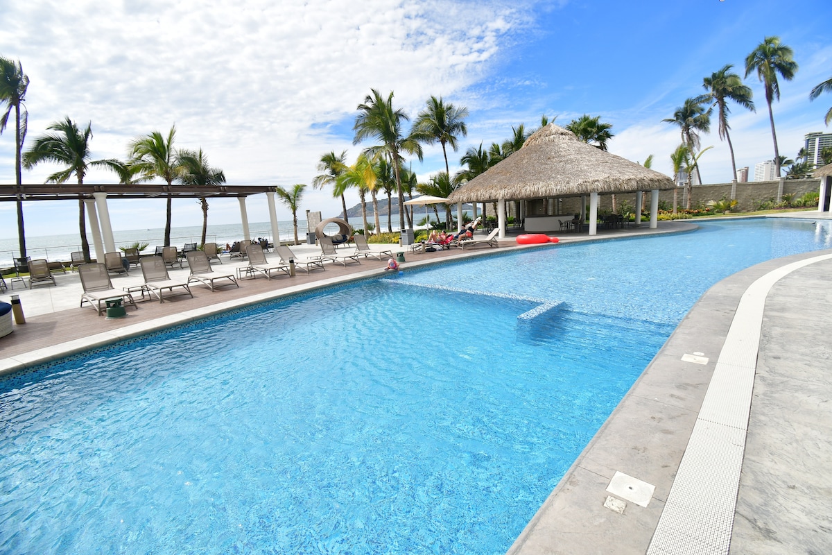 A large outdoor pool is surrounded by palm trees, with a thatched-roof gazebo in the background. Lounge chairs are arranged along the pool deck, offering a place to relax. The serene water reflects a clear sky, creating a tranquil atmosphere.
