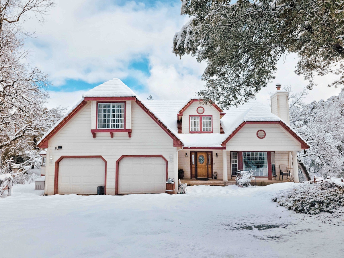 A two-story mountain home is portrayed under a blanket of snow, highlighted by a light-colored exterior and red accents. Icicles hang from the roof, and the driveway is partially visible, leading to the attached garages on either side. Pine trees surround the property.