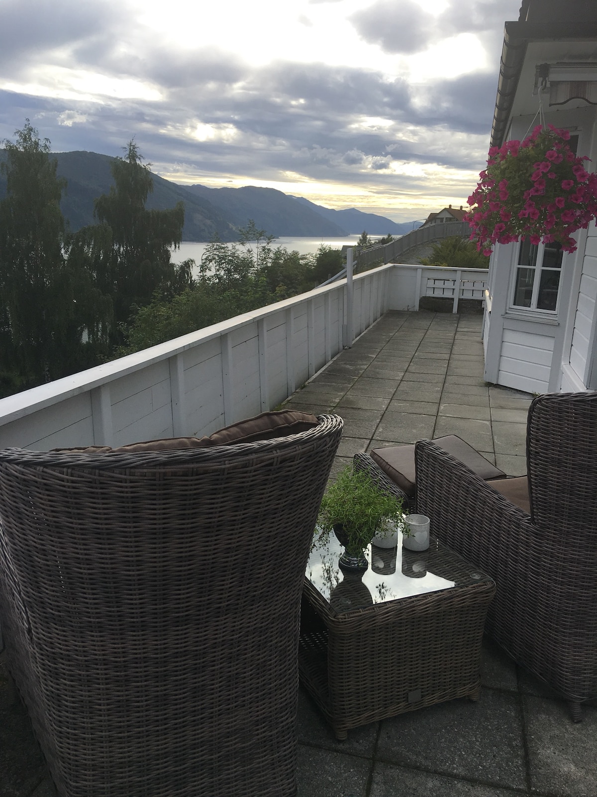 A spacious terrace is presented with two wicker chairs facing the scenic view of mountains and a fjord. A small table, adorned with decorative items, is positioned between the chairs. Flower baskets hang from the railing, adding a touch of color to the serene outdoor space.