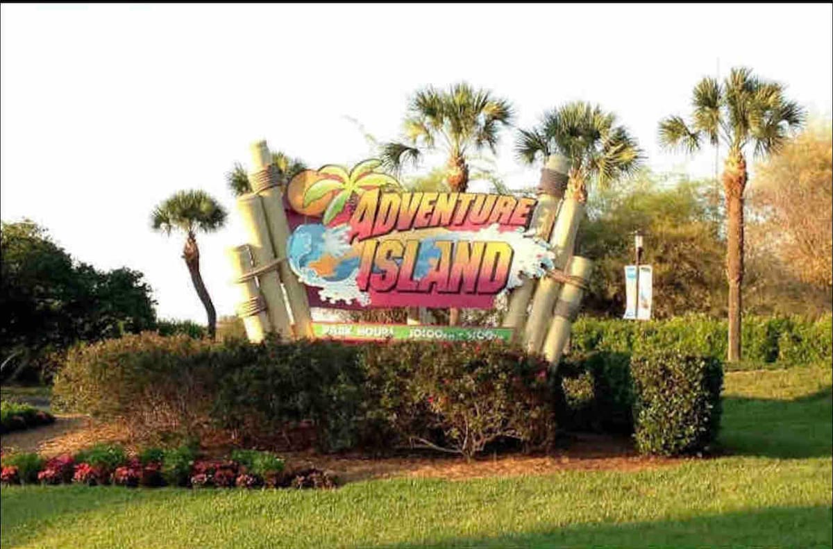 The entrance of Adventure Island is prominently displayed, featuring the vibrant sign adorned with tropical designs. Surrounding the sign are neatly maintained shrubs and colorful flower beds, with palm trees and lush greenery providing a scenic backdrop.
