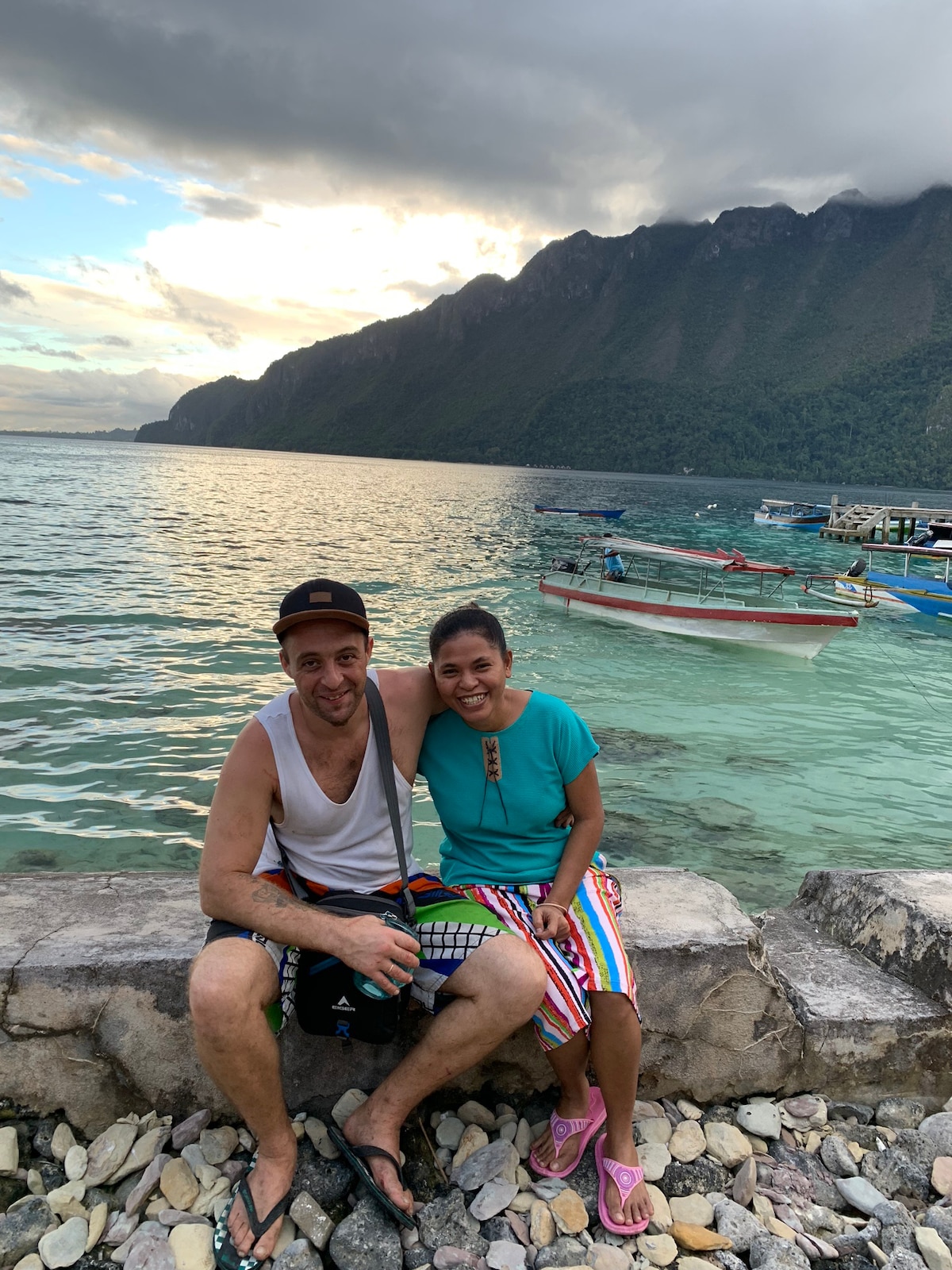 A serene waterfront scene captures two individuals seated on a stone edge, overlooking clear waters. Small fishing boats are visible in the background, while lush mountains rise majestically in the distance under a partly cloudy sky.