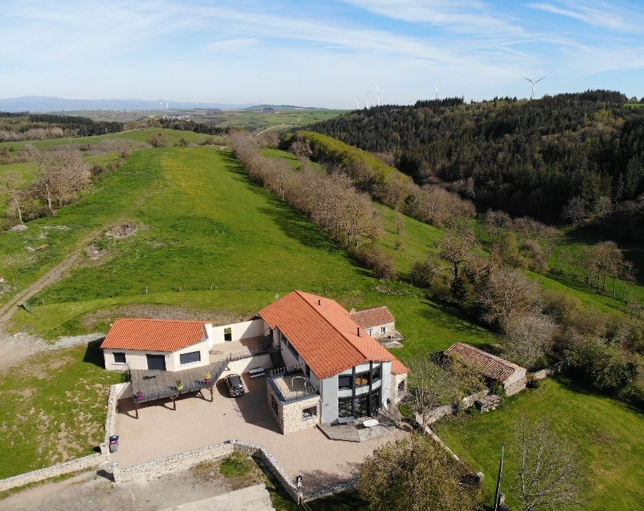 An aerial view of a recently built gîte is presented, showcasing a spacious property surrounded by lush greenery. The terracotta-tiled roof complements the natural landscape, which features fields and distant hills. Wind turbines can be seen on the horizon, contributing to the serene rural atmosphere.