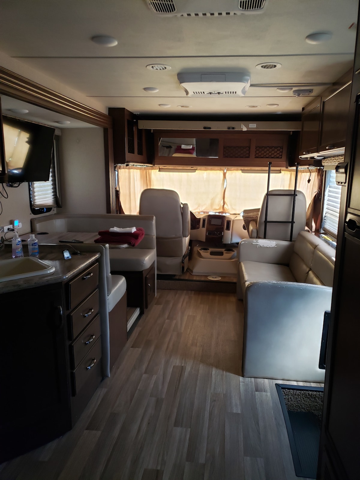 An interior view captures a well-organized RV space featuring a compact kitchen area with cabinetry, a seating arrangement with a light-colored couch, and a driver's seat visible. Soft natural light filters through the front windows, illuminating the warm wood tones and addressing the overall functionality of the space.