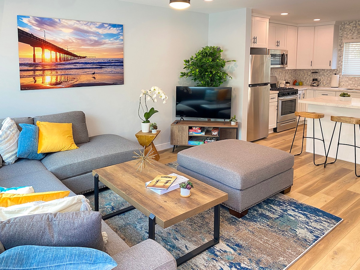 A modern living area features a gray sectional sofa with colorful accent pillows, a coffee table, and a stylish rug. A flat-screen TV is positioned across from the seating, while a poster of a pier at sunset decorates the wall. A dining area with bar stools is visible in the background.