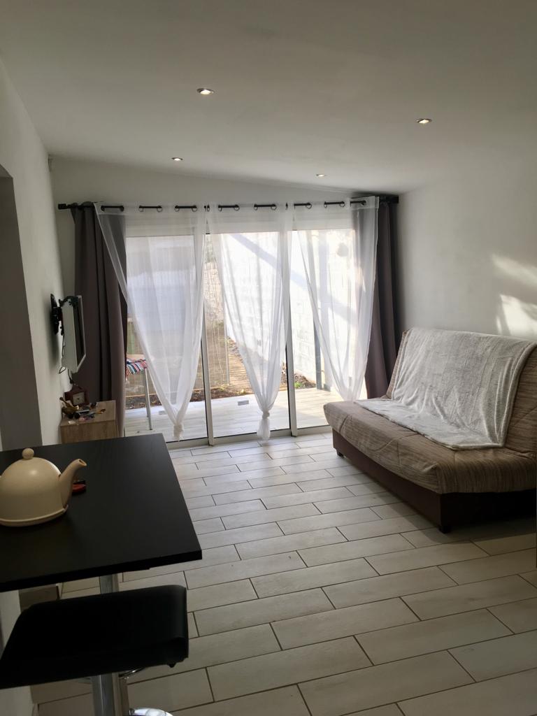 A living area is depicted, featuring a sofa bed and a small table with a kettle. Natural light is filtered through sheer curtains hanging above sliding glass doors that lead to an outdoor space. Light-colored tiles cover the floor, enhancing the airy atmosphere.
