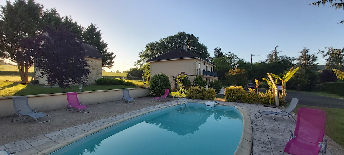 A tranquil outdoor space features a shared swimming pool surrounded by a sun terrace. Colorful lounge chairs are arranged around the pool, with lush greenery and trees in the background. The early morning light casts a serene glow over the area, enhancing the peaceful atmosphere.