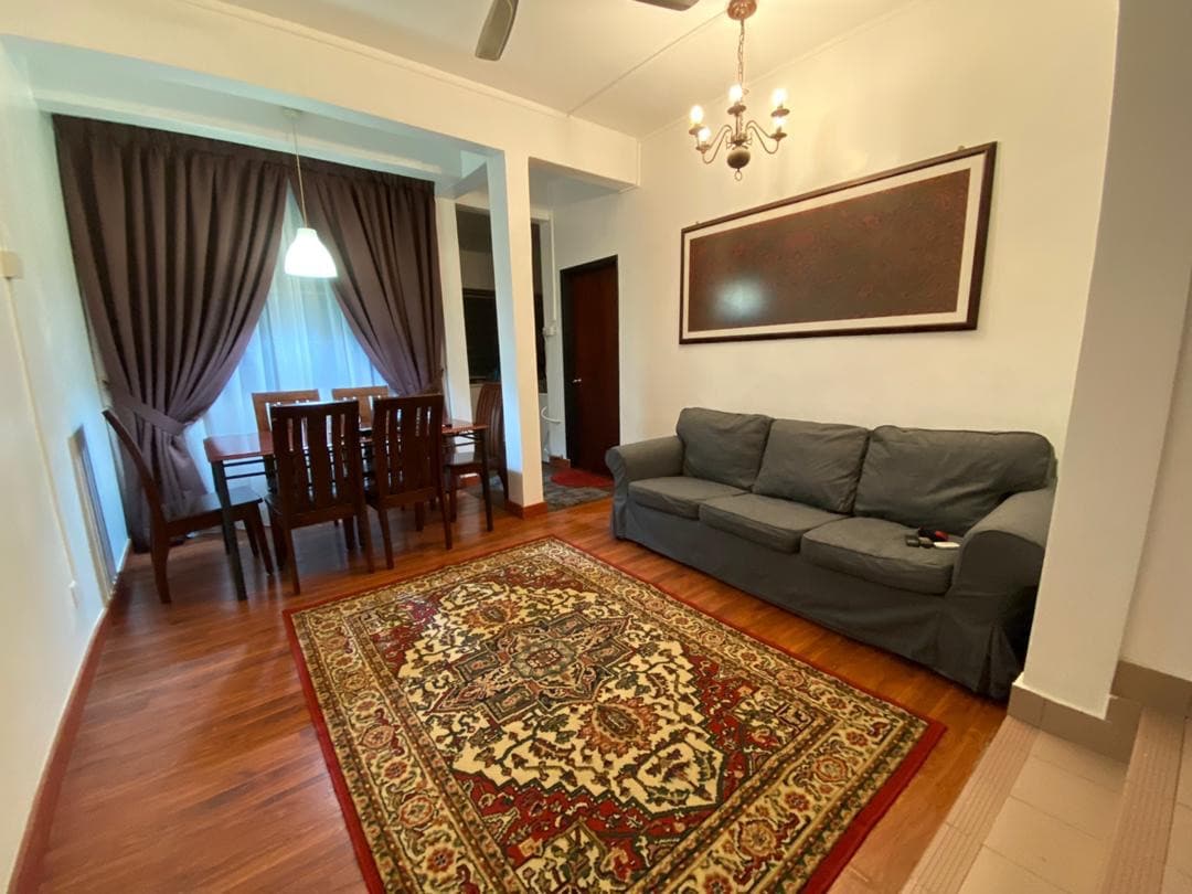 A living area features a comfortable gray sofa positioned on a patterned area rug. A dining table with wooden chairs is arranged nearby. Soft curtains frame a window, and a chandelier adds a touch of elegance to the space.