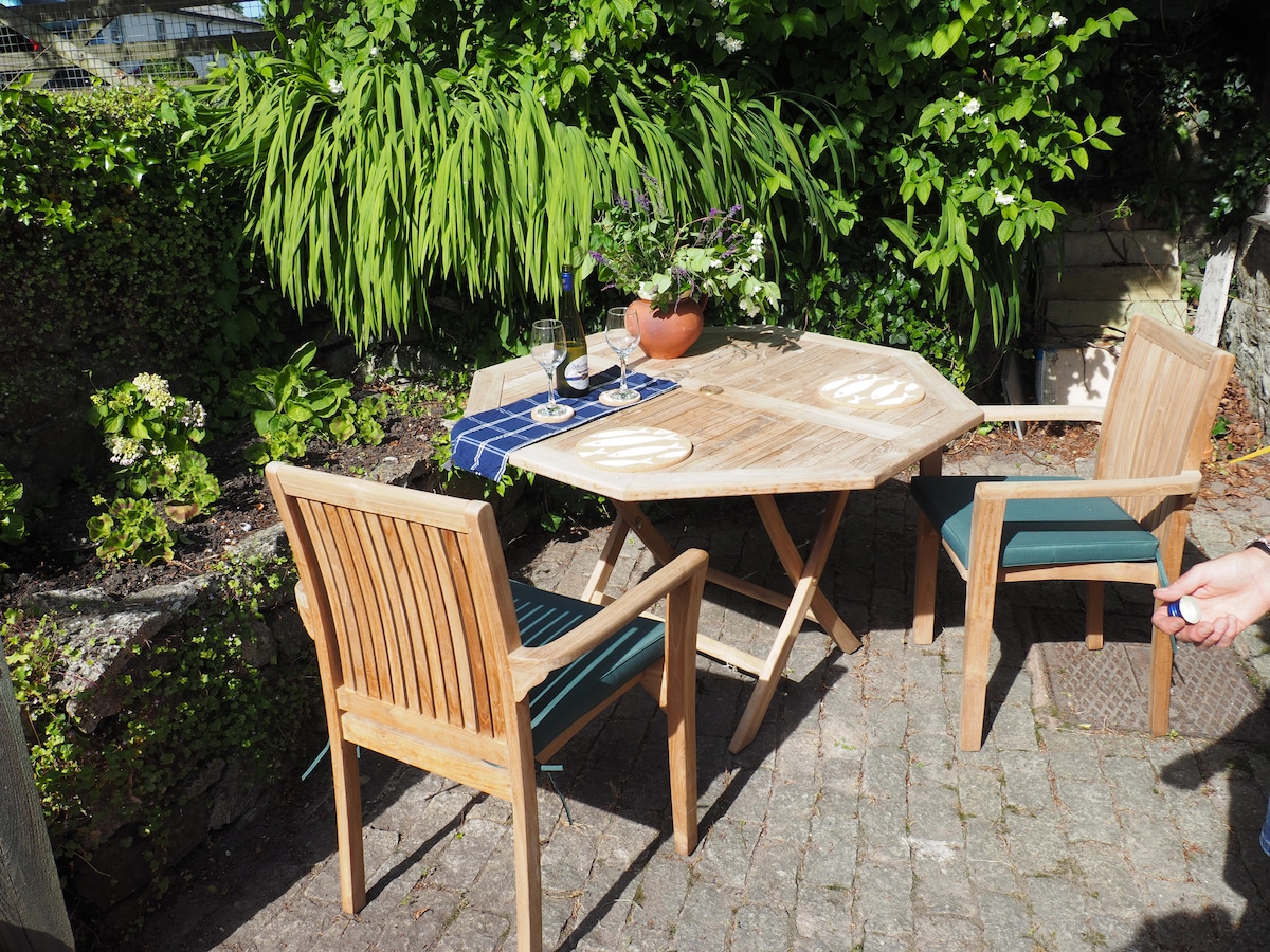 An outdoor dining area is set with a wooden table and two matching chairs. A tablecloth is adorned with a blue checkered design, and a small plant is placed at the center. Surrounding greenery adds a touch of nature to the space.