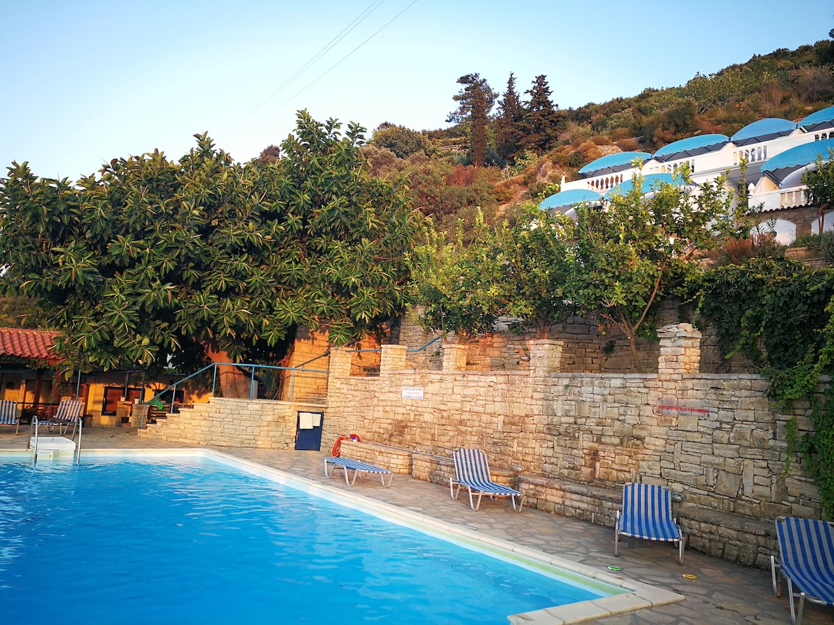 A serene swimming pool is surrounded by stone walls and lush greenery. A row of lounge chairs with blue and white striped fabric are positioned by the water. In the background, terraced buildings with blue rooftops can be seen, complementing the scenic hillside.