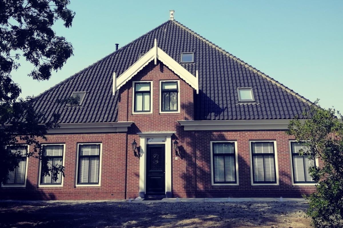 A traditional Dutch Stolp farmhouse is presented, showcasing a sloped tiled roof and a brick facade with decorative white trim. Windows adorn the structure evenly, allowing ample natural light. A shaded area in front provides a sense of space, complemented by surrounding greenery.